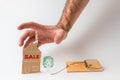 Bank investments and mortgages. Male hand reaching for the cardboard house, lying next to the trap. Text SALE. White background