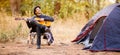 Banner format image of smiling young man in black hat sitting near touristic tent and playing guitar in forest Royalty Free Stock Photo