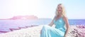 Banner girl blonde sitting on the rocks by the sea blue dress sky lighthouse sun beach horizon summer. Royalty Free Stock Photo