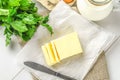 A bar of butter is cut into pieces on a wooden board with a knife, surrounded by milk, eggs and parsley on a white table. Ingredie