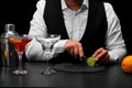 A bartender cuts a sappy green lime with a knife, a bar counter with glasses, oranges on a black background. Royalty Free Stock Photo