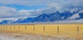 Barbed wire fence runs around a golden pasture under the snowy Rocky mountains.