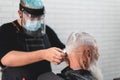 Barber cuts a senior hipster man hair with a razor and clipper while using a face shield - Focus on customer