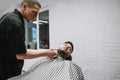Barber with hair clipper in the hands makes a hairstyle in the beard of the client, lying on a chair and looking away with his