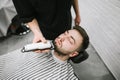 Barber with hair clipper in the hands makes a hairstyle in the beard of the client, lying on a chair. Male hairdresser doing