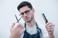 Barber holds the comb and scissors on his arms outstretched against the background of a white wall.