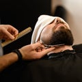 A barber stylist trims the beard of a Caucasian man, whose face is covered with towel, with scissors Royalty Free Stock Photo