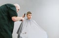 Barber trimming young man`s hair on a gray background, customer looking into camera with serious face. Creating stylish male