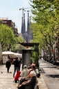 Barcelona city street scene, people relaxing on the benches. BasÃÂ­lica de la Sagrada Familia by Antoni Gaudi in the distance