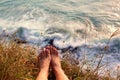 Bare feet of a female person laying on grass above swirling water of stormy ocean at sunset. Freedom during travel trip