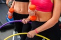 Bare female belly with dumbbells and hoop at gym.