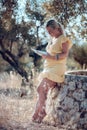 A barefoot blonde woman with glasses in a yellow summer dress is reading a book in the shade of olive trees