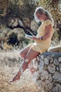 A barefoot blonde woman with glasses in a yellow summer dress is reading a book in the shade of olive trees