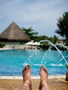 Barefoot feet of a man taking a sun bath in a pool with fountains nd a hut on the background