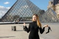 Barefoot woman standing near Louvre and glass pyramind in black dress in Paris with shoe in hand.