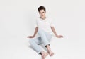 A barefoot young male model in jeans and a white T-shirt sits on the floor.