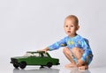 Barefooted baby boy toddler in blue one-piece bodysuit romper with long sleeves sits squatting plays with big toy car Royalty Free Stock Photo