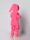 Barefooted baby girl in pink warm comfortable jumpsuit with hood on and bunny ears stands back to camera