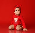 Barefooted calm infant baby girl toddler in red bodysuit at Christmas New Year sits on floor looking at camera