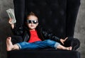 Impudent kid boy millionaire in leather jacket and red t-shirt is sitting lounging in armchair holding a bundle of cash