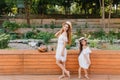 Barefooted young woman and little girl standing back to back in front of flowerbed. Outdoor full-length portrait of