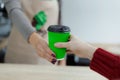 Barista in apron is giving hot coffee in green takeaway paper cup to customer. Coffee take away at cafe shop