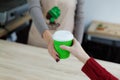 Barista in apron is giving hot coffee in green takeaway paper cup to customer. Coffee take away at cafe shop