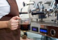 Barista dressed in brown apron holding a measuring cup fill with hot coffee. Morning atmosphere in a coffee shop