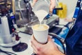 Barista using coffee machine preparing fresh coffee and pouring milk to espresso coffee make latte art into white paper cup at cof