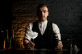 Barman holds transparent glass full of ice and shaker which stands on bar counter