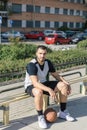 Basketball player sitting next to his basketball ball on an urban court, waiting to go out to play Royalty Free Stock Photo