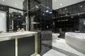 Bathroom with white porcelain sink above chest of drawers and beveled mirror above wall sconce in a room with black tiles and a Royalty Free Stock Photo