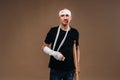 A battered man with a bandaged head and a cast on his arm stands on crutches on a gray background Royalty Free Stock Photo