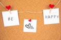 Be happy handwriting black lettering on 3 white papers pinned with 2 big red heart pegs and one small red heart pegs on cork board