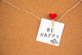 Be happy lettering with heart on white paper fastened to the right with a heart-pin on a cork board