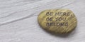 Be here, be you, belong symbol. Beautiful stone with words `Be here, be you, belong` on beautiful white wooden background.