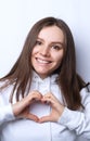 Charming young girlfriend confesses in love, shows heart gesture, smiles broadly, has romantic feelings