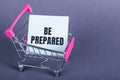 BE PREPARED is message on white card in a shoppping cart on violet background. top view