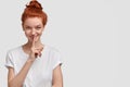 Be quiet. Cunning intriguing red haired woman has hair bun, keeps fore finger over lips, asks to be speechless, looks Royalty Free Stock Photo