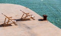 Beach deck chairs facing the sun at turquoise sea and rusty bollard