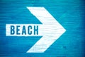 Beach text sign written on a white directional arrow pointing towards right manually painted on a blue wooden signboard