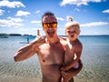 Beach vacation: A guy in sunglasses on the sea shore holds a child in the arms and shows the symbol of Aloha. Portrait. Royalty Free Stock Photo