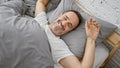 Beaming middle-aged man with grey hair joyfully stretching arms waking up in his cozy bedroom bed, relishing the comfort of a Royalty Free Stock Photo