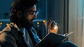 Bearded adult man in glasses at night home living room with flashlight shines on book reads preparing for exams reading Royalty Free Stock Photo