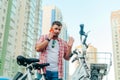 Bearded adult stands by a bicycle against the backdrop of buildings and the sky, talking on a smartphone with a serious face. Royalty Free Stock Photo