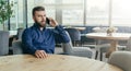 Bearded attractive businessman in blue shirt is sitting at wooden round table in restaurant and talking on cell phone. Royalty Free Stock Photo