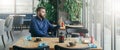 Bearded attractive businessman in blue shirt is sitting at wooden table near window in restaurant and holding smartphone Royalty Free Stock Photo