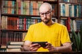 A bearded bald man with glasses uses a smartphone with a frown. Bookshelves in the background. The concept of a social network,