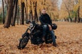 Bearded biker poses on own motorcycle, holds helmet, rides motorbike, poses outdoor in park, ground covered with fallen leaves.