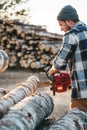 Bearded brutal lumberjack wearing plaid shirt sawing tree with chainsaw for work on sawmill. Sawdust fly apart